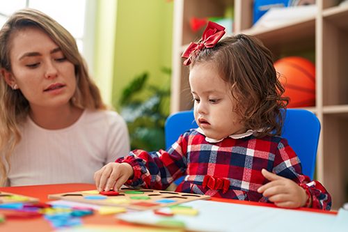 An education assistant supervises a young girl as she works on a colorful puzzle