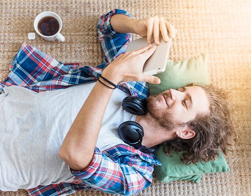 A scruffy hipster lays on the floor enamored with his tablet 